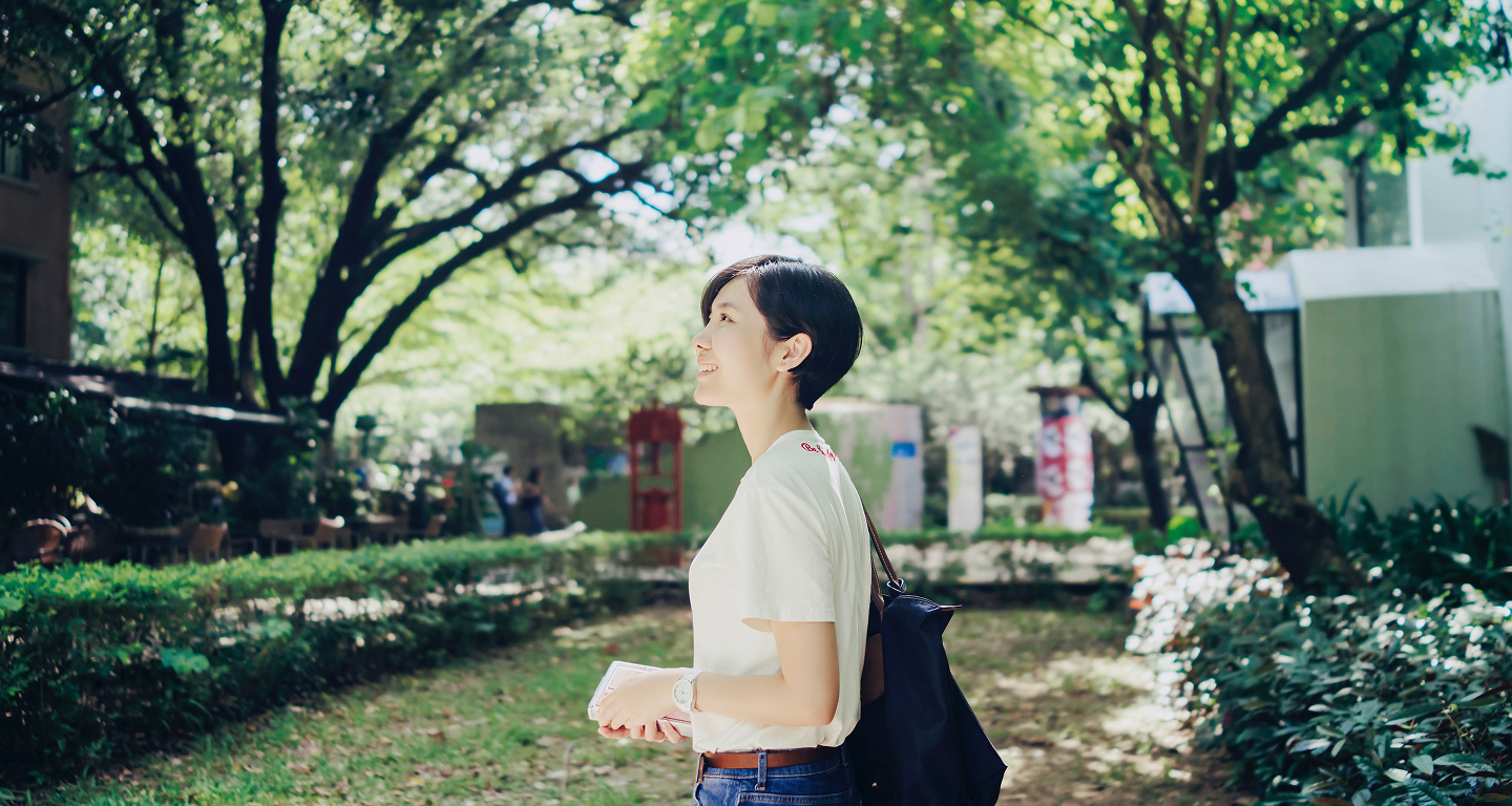 緑豊かな木々を背景に、白いTシャツを着て笑顔で立つ女性の横顔